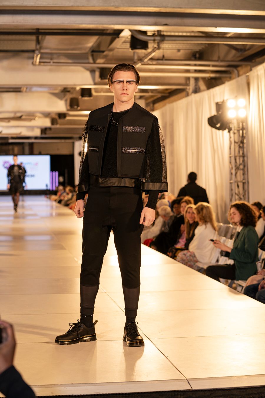 Model showcases black tactical streetwear ensemble on illuminated white runway during fashion show.