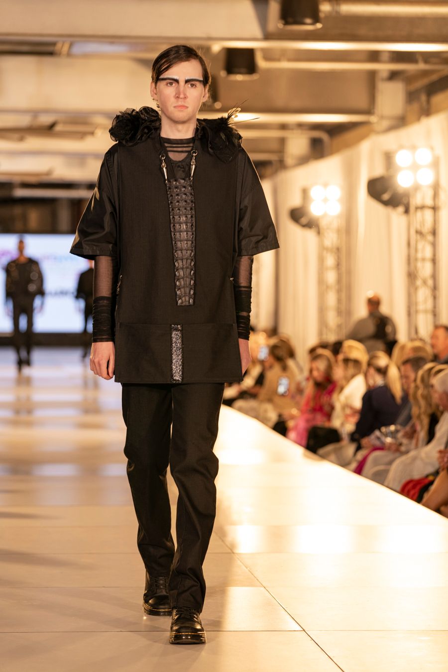 Model presents all-black casual outfit with graphic t-shirt on white catwalk during indoor fashion event.