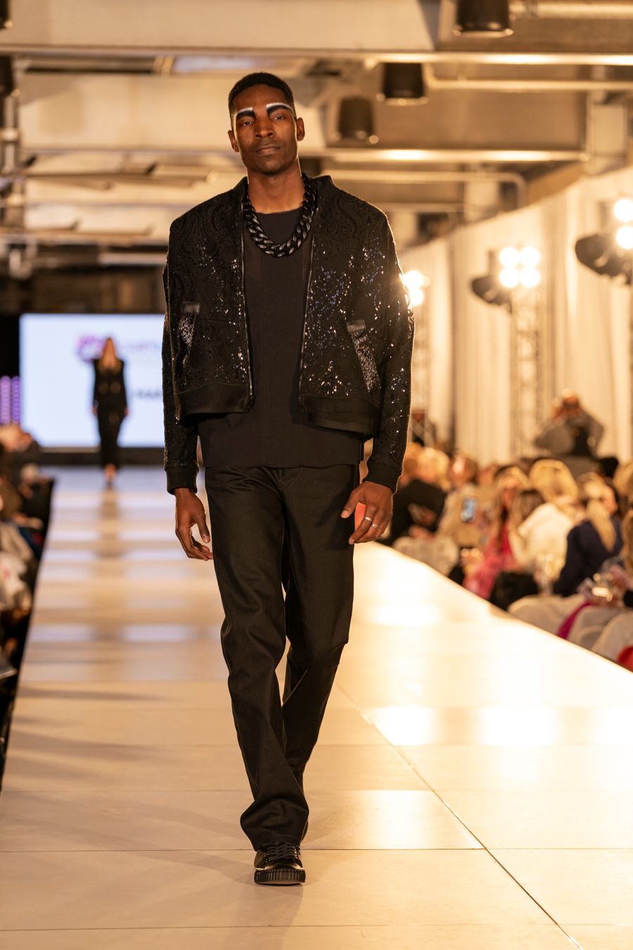 Model walks down white runway in black outfit during plus size fashion show at indoor venue.