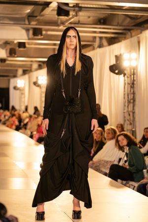 Fashion model walks runway in dramatic black hooded maxi dress with flowing silhouette at indoor fashion show.