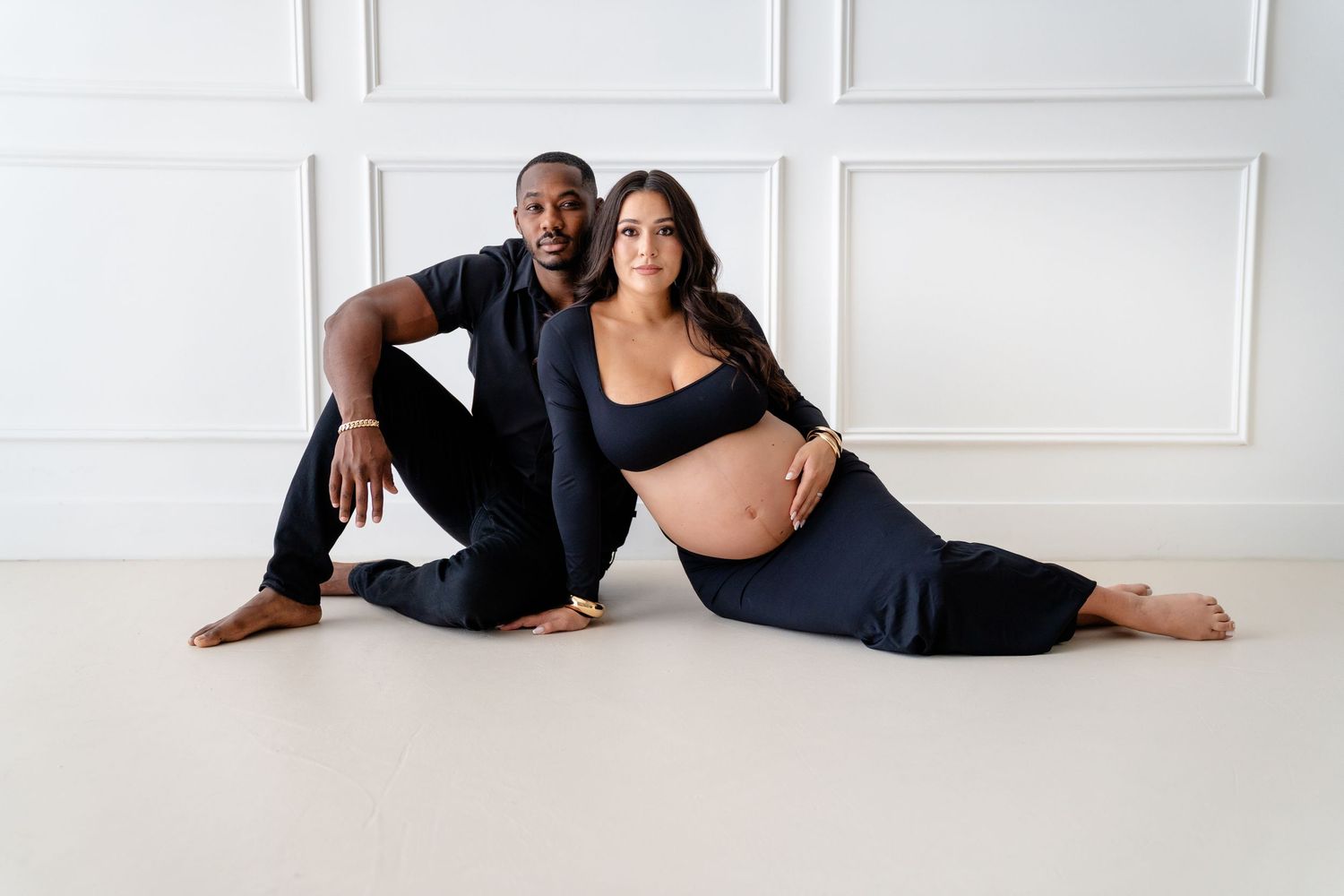 A couple poses together on a white floor during a maternity photoshoot, with the woman wearing black athletic wear.