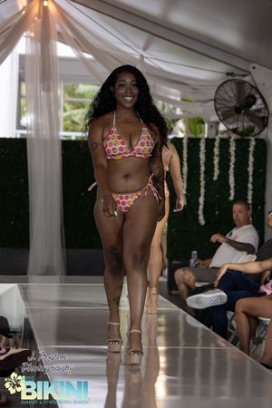 Model walks runway in floral print bikini at outdoor fashion show.