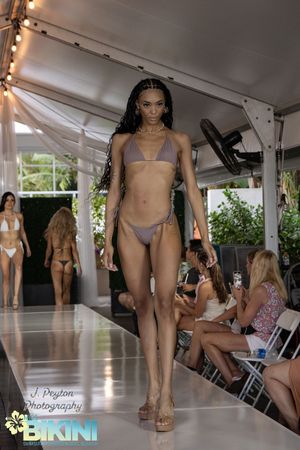 Models showcase purple and neutral toned swimwear on a covered runway.