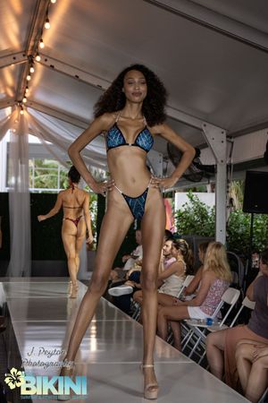 Models walk the runway showcasing swimwear designs during a fashion show under a white tent structure.