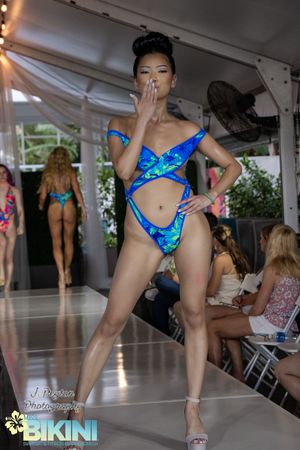 Models display blue patterned swimwear during a summer fashion show under a white canopy.