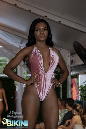 Model walks runway in pink velvet plunging swimsuit at outdoor fashion show.