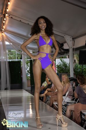 Models walk the runway in purple swimwear at a fashion show under a white tent.