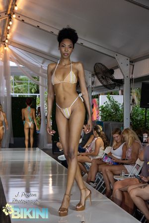 Model walks runway in white string bikini at summer fashion show with audience seated along catwalk.