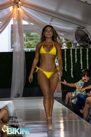 Models walk the runway in bright yellow bikinis during a summer fashion show under a white tent.