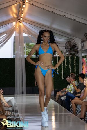Fashion models showcase blue bikinis and swimsuits on an illuminated runway with audience seated alongside.