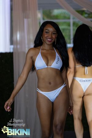 Models walk the runway in white bikinis at a summer fashion show under a white tent.