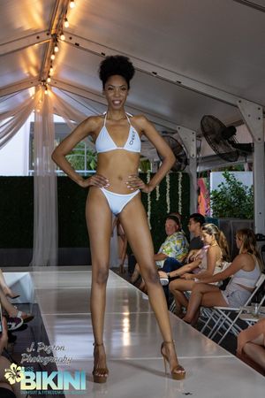 Fashion models showcase white swimwear designs on a covered outdoor runway with audience seated along sides.