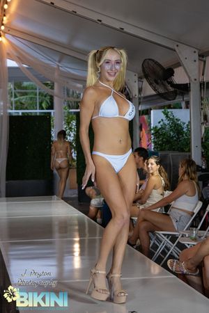 Models walking a runway in white bikinis and high heels during a fashion show event under a covered venue.