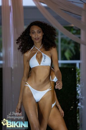 Fashion models walk the runway in white swimwear at a beachwear fashion show under a white tent.