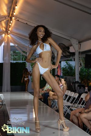 Models walk the runway in white bikinis during a fashion show under a white tent with folding chairs.