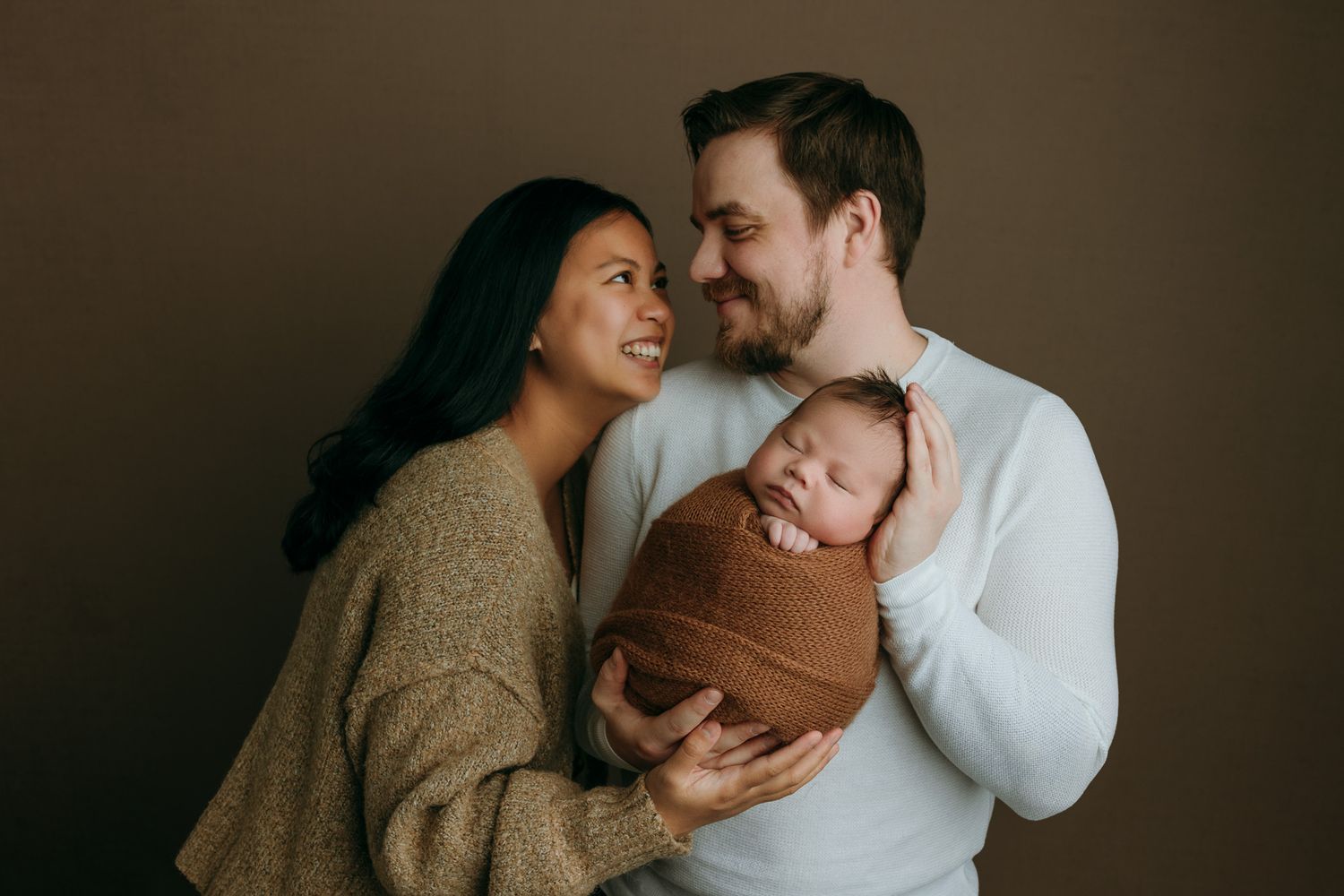 A couple in cozy sweaters share a tender moment while holding their newborn baby wrapped in brown fabric.