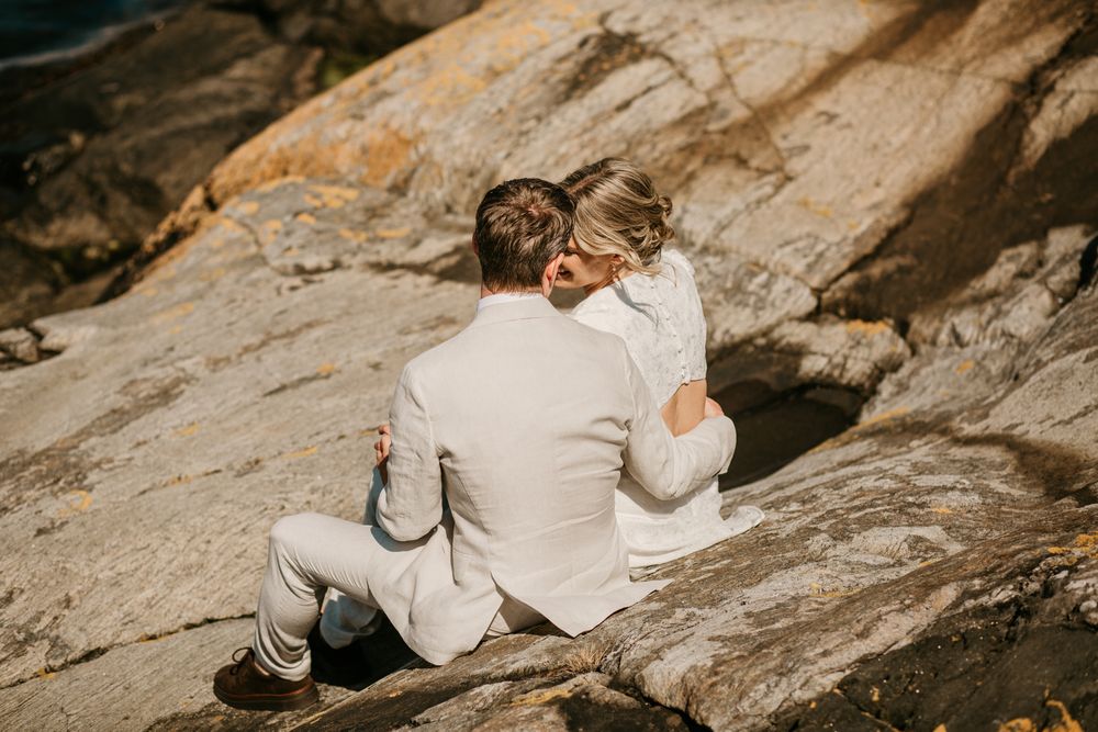Two people in white outfits sit close together on rocky terrain during a sunny outdoor photo session.