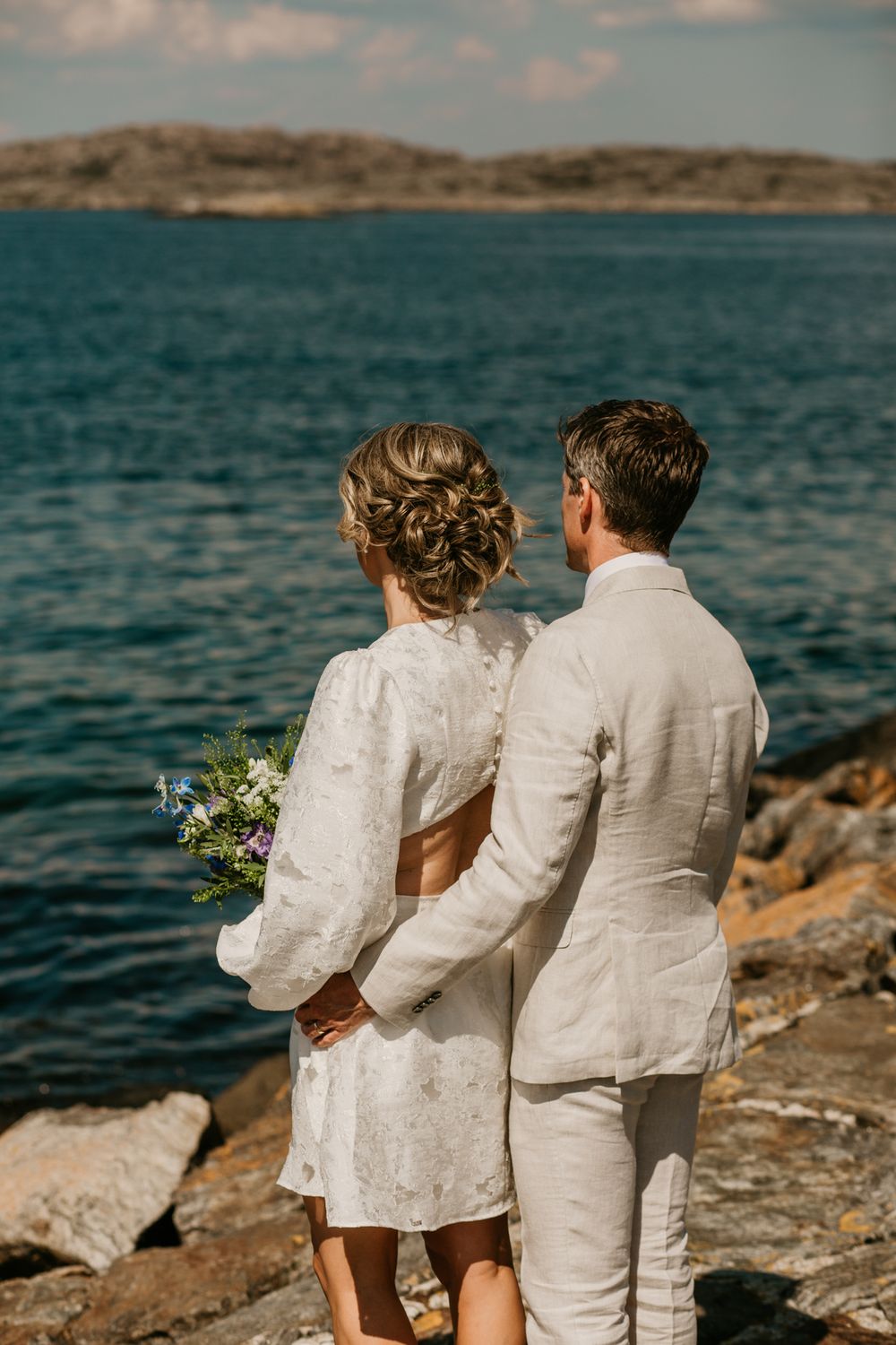 A couple in white attire embraces on rocky coastal cliffs overlooking turquoise waters on their wedding day.