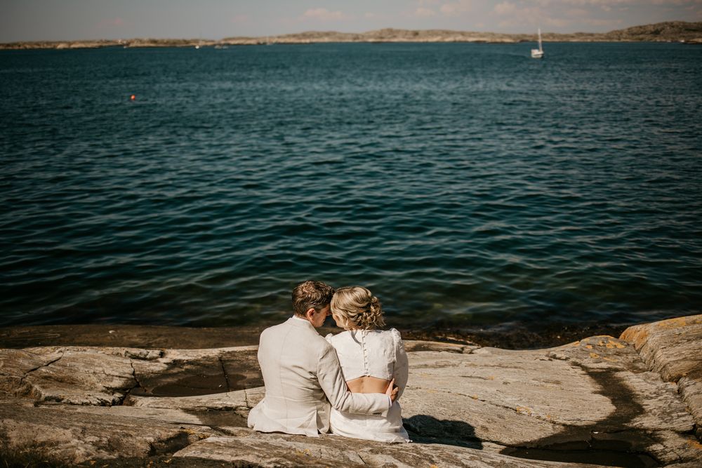 Couple in wedding attire sits on rocky shoreline overlooking tranquil blue waters and distant mountains.