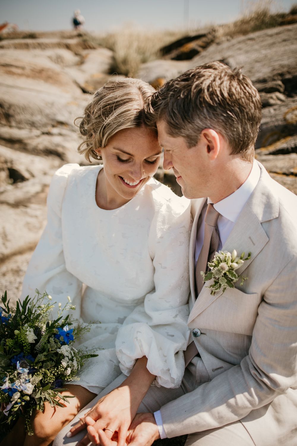 A romantic couple in white wedding attire share intimate moments on a rocky beach during their elopement photoshoot.