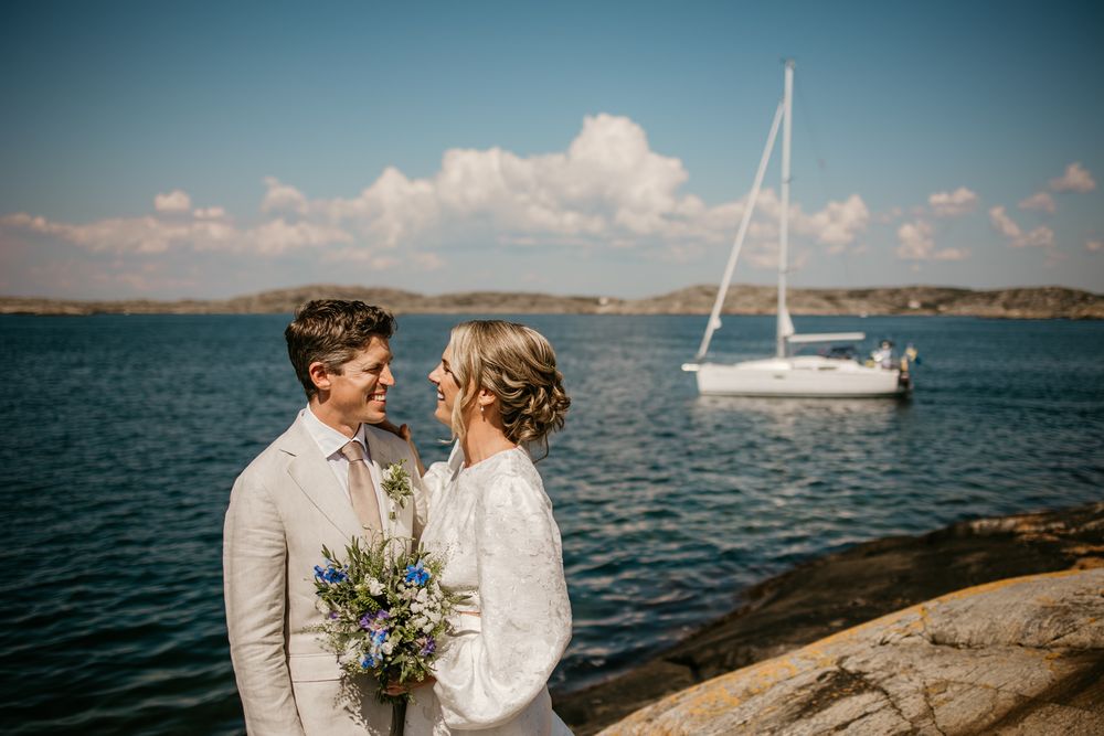 A romantic seaside wedding moment with a sailboat in the background as the couple stands on rocky shores.