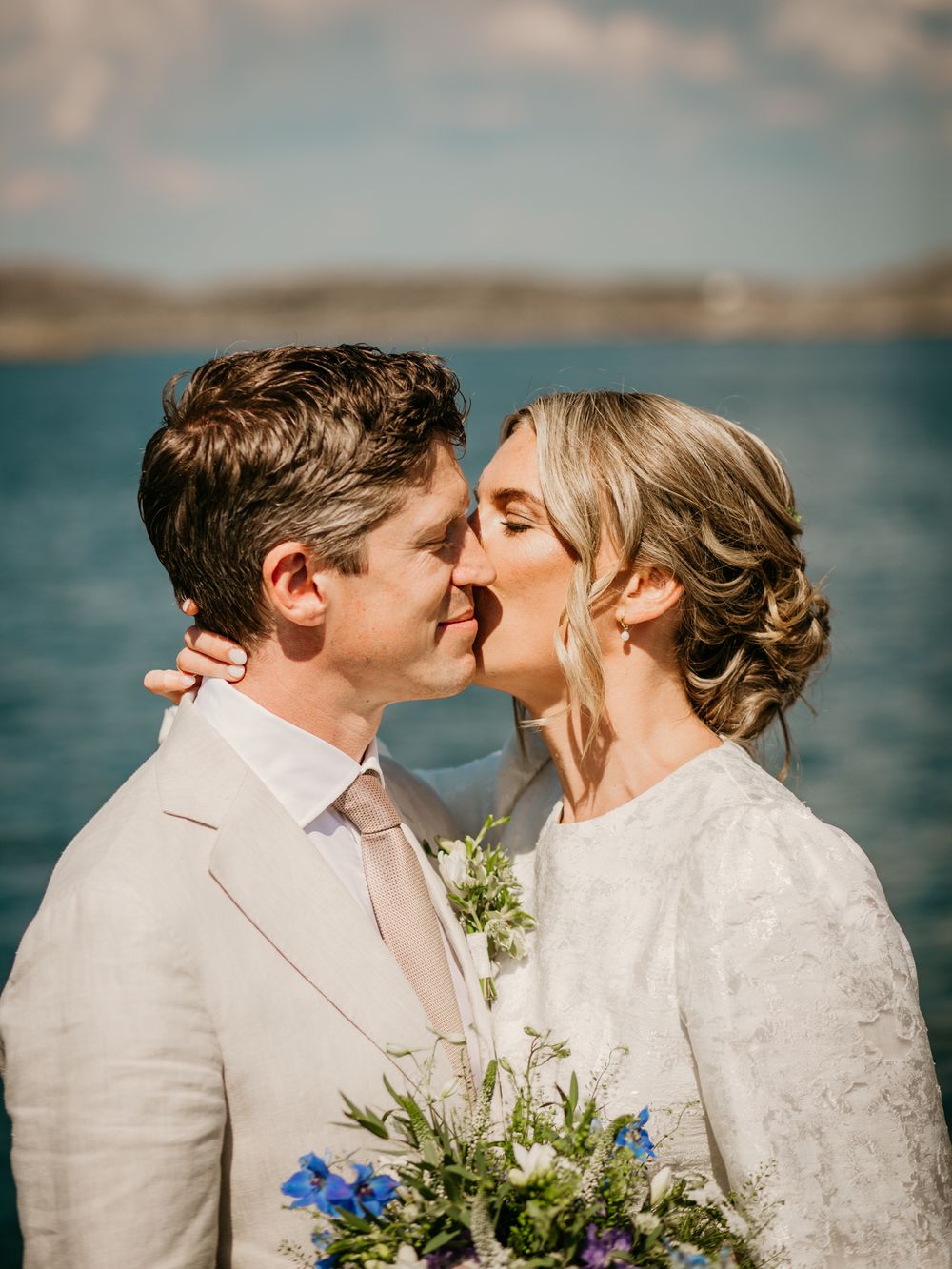 A romantic lakeside wedding moment with a couple in white attire sharing a tender kiss against a scenic water backdrop.