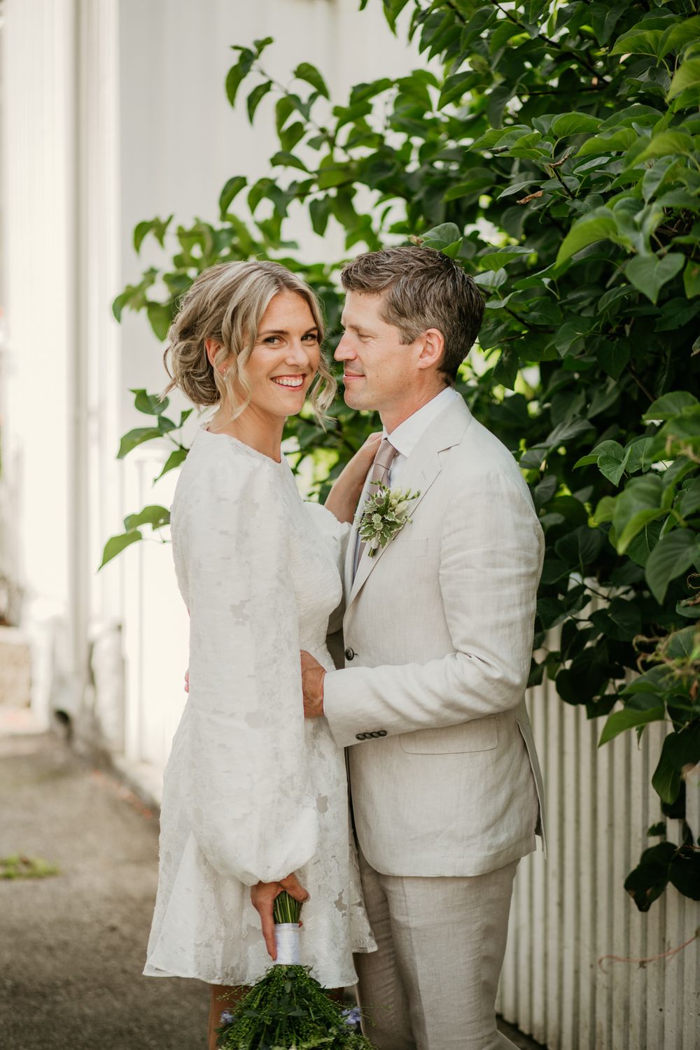 A happy couple shares a romantic moment in their white wedding attire on a porch surrounded by green foliage.