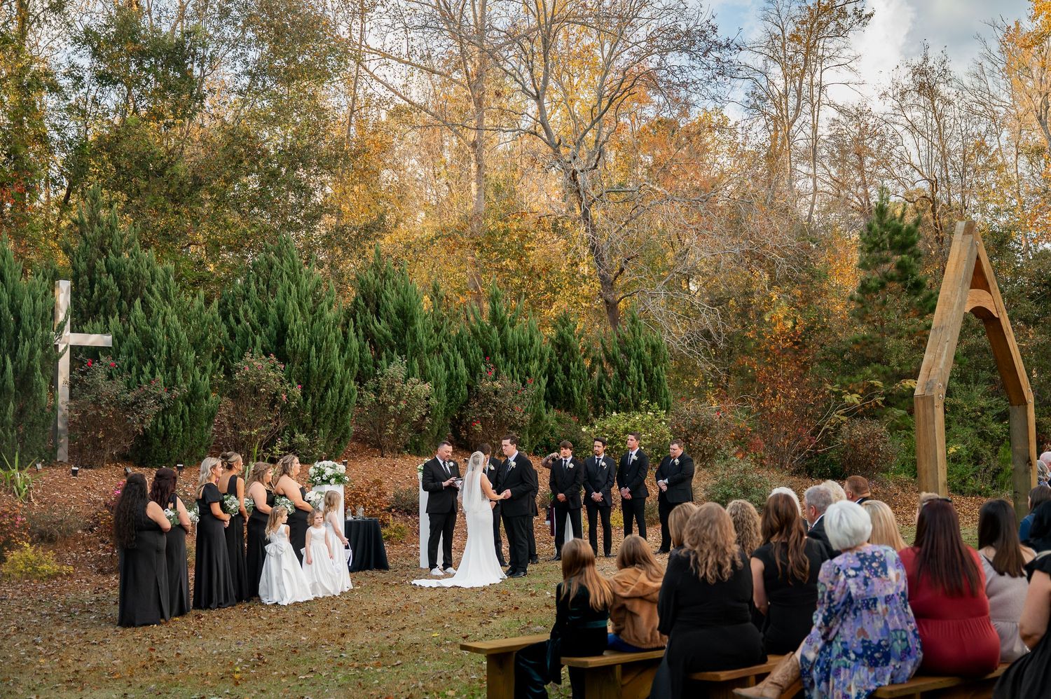 Wedding ceremony taking place outdoors in autumn with wooden arch and guests gathered on lawn.