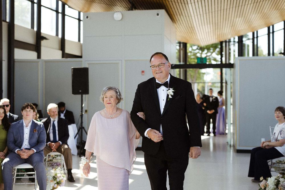 Wedding guests gather in a modern reception venue with wood paneled ceiling and large windows.