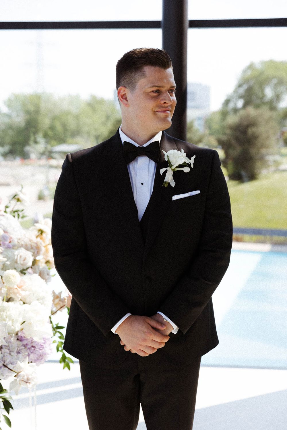 Groom in classic black tuxedo smiling during outdoor wedding ceremony by waterfront.
