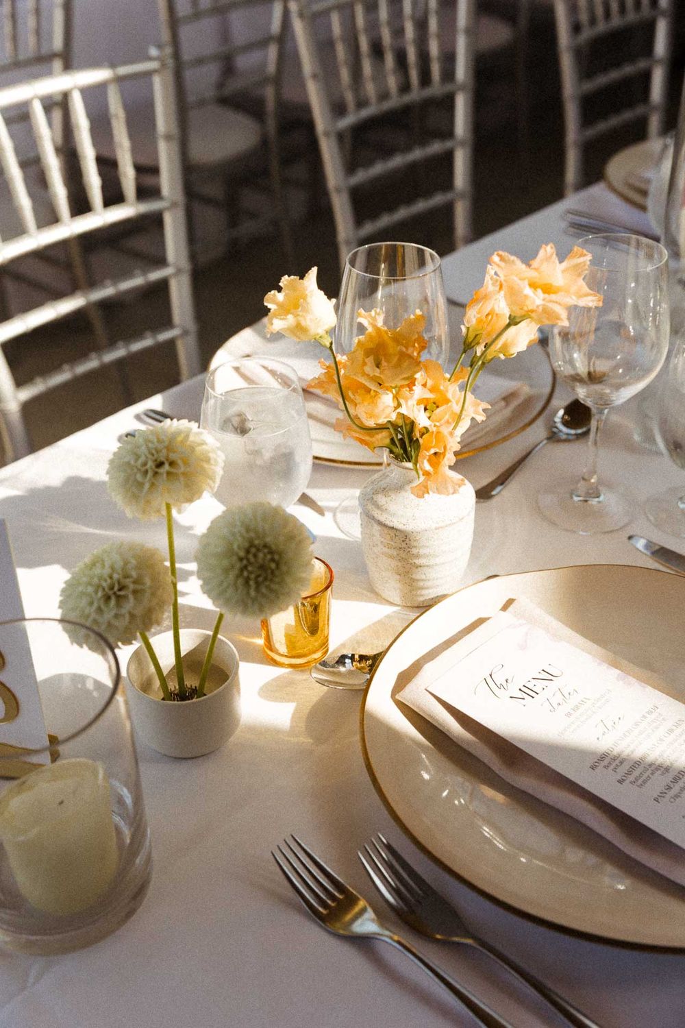 Elegant wedding reception table settings with white floral centerpieces and gold accents in natural sunlight.