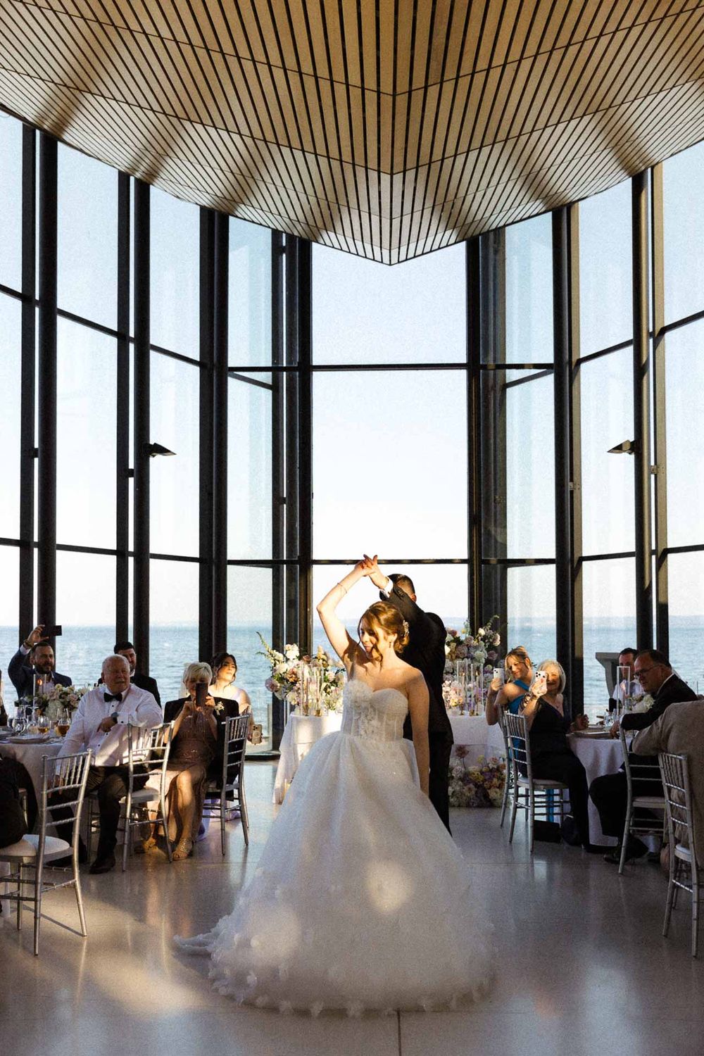 Wedding reception in a modern venue with floor-to-ceiling windows and wooden slat ceiling.
