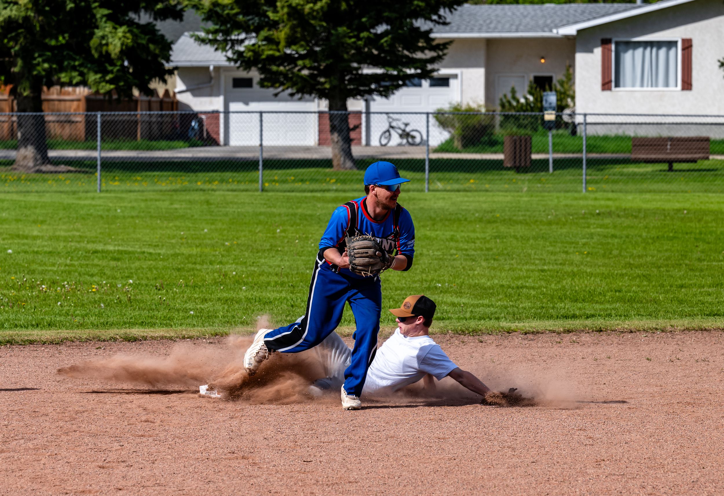 Baseball player sliding into base during game on dirt infield with green grass and buildings in background.