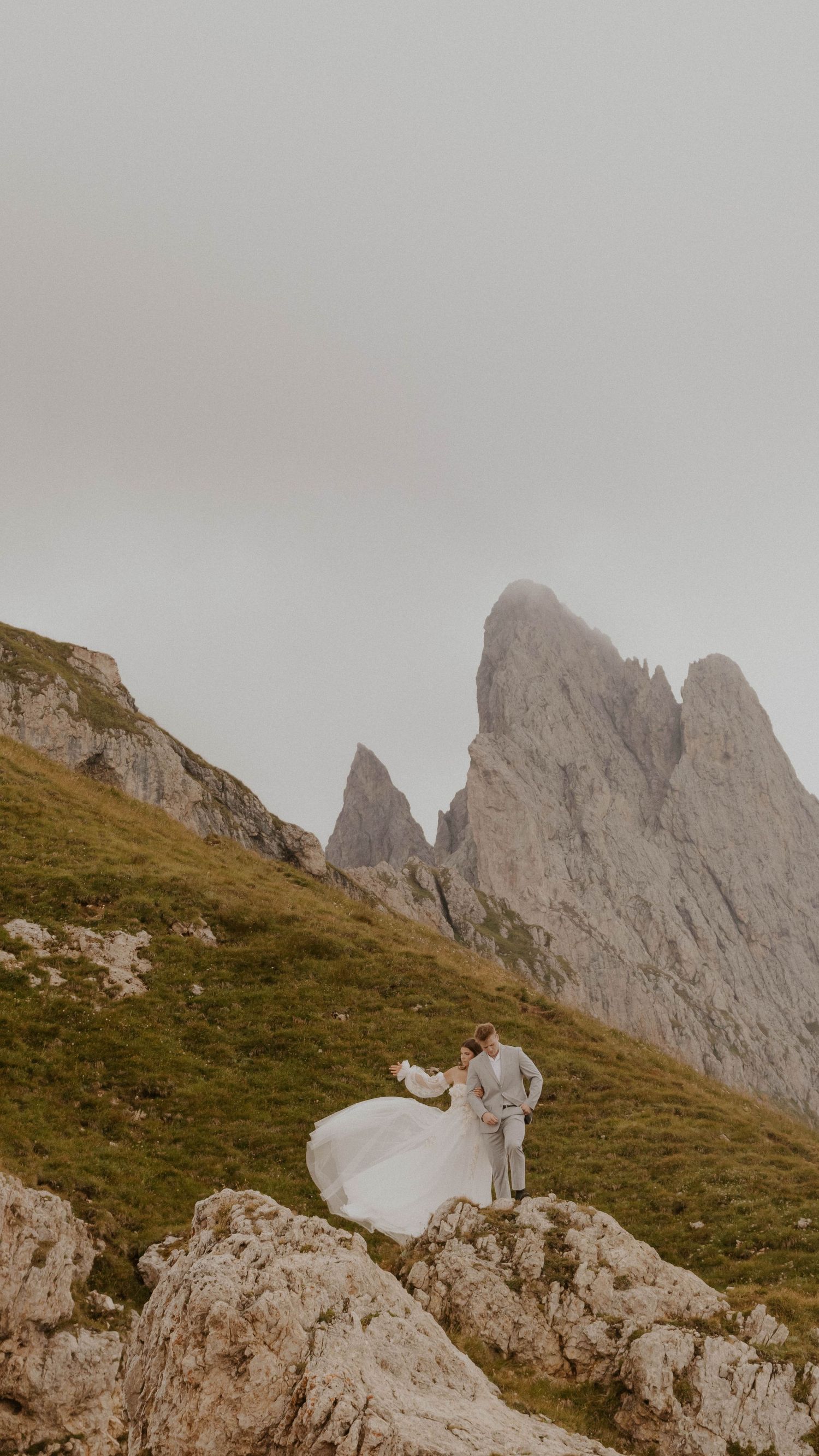 A couple in wedding attire stands on a rocky mountain peak with dramatic Dolomite peaks rising in the misty background.