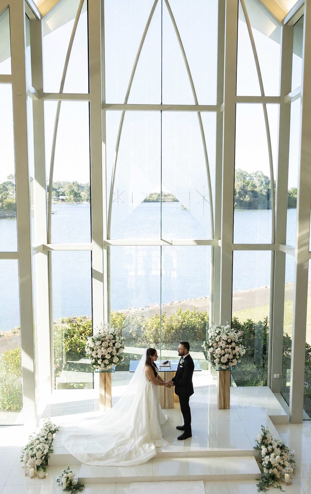 Bride and groom exchange vows at altar beneath soaring arched windows overlooking a serene waterfront chapel.