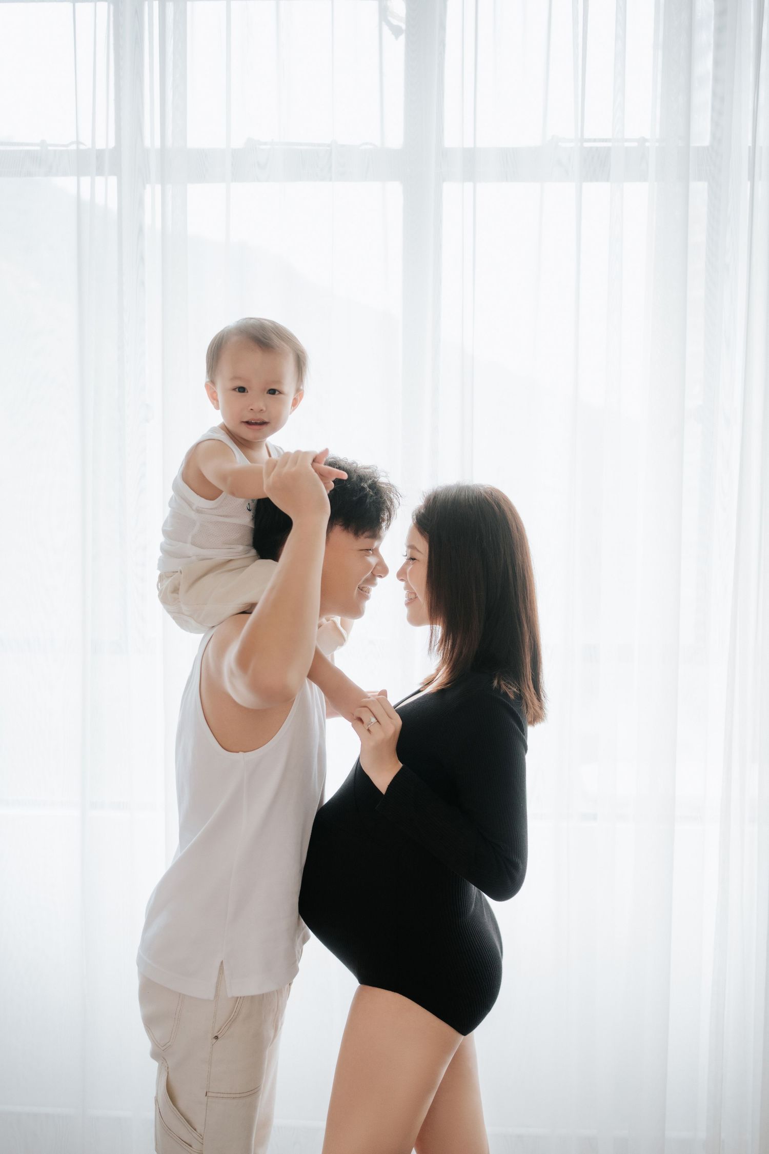 A couple shares a tender moment with their baby in front of sheer white curtains in a bright, minimalist setting.