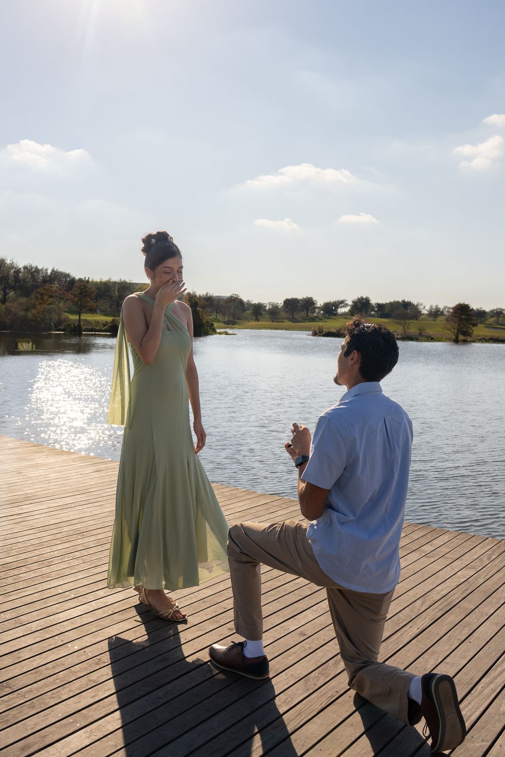 A romantic proposal scene unfolds on a wooden dock by a serene lake on a sunny afternoon.