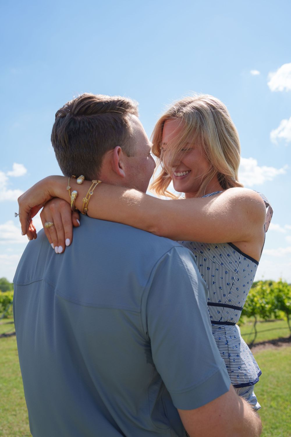 A couple embracing and smiling at each other outdoors in a vineyard under a bright blue sky.
