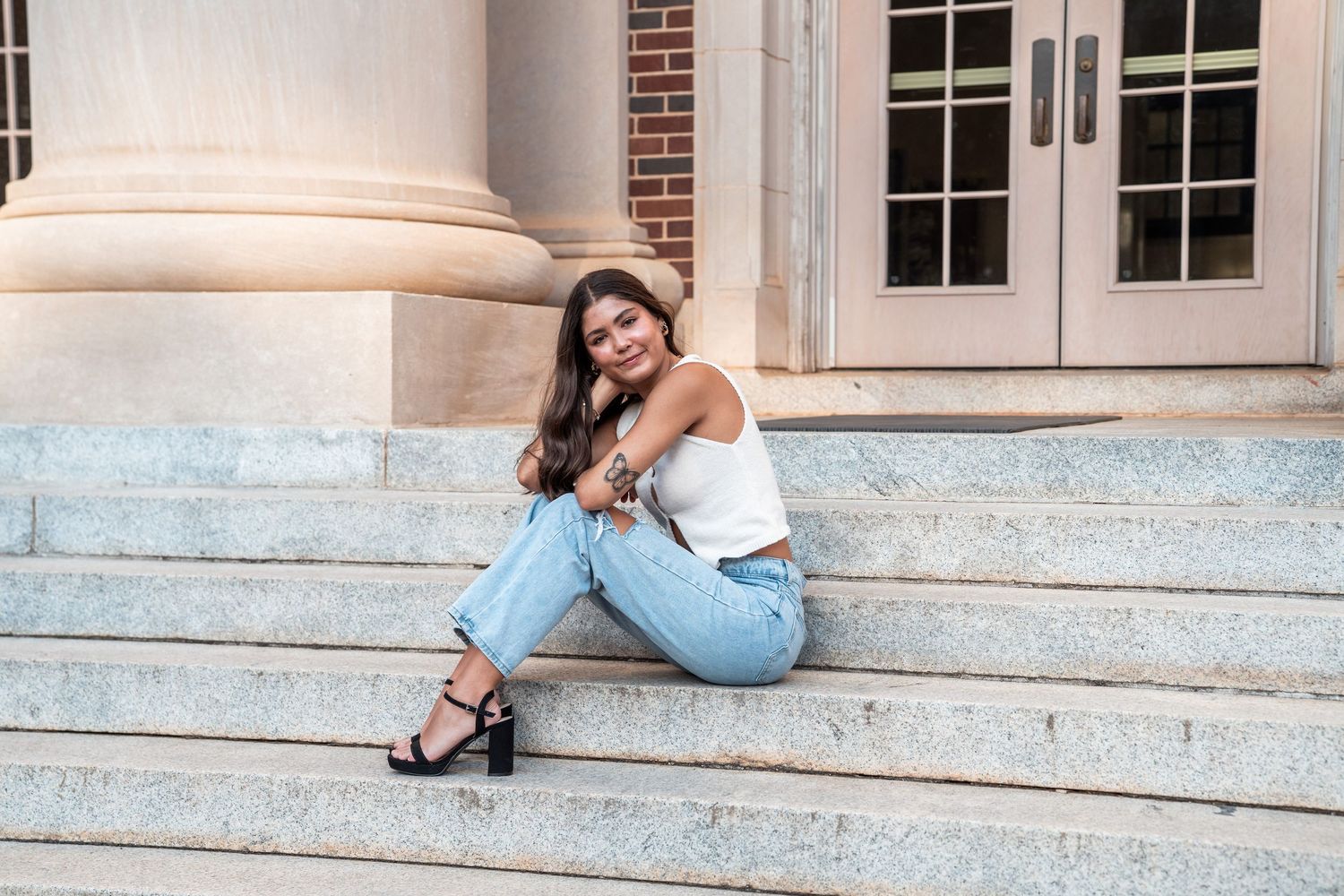A person in a white tank top and light blue jeans sits casually on stone steps near a brick building entrance.