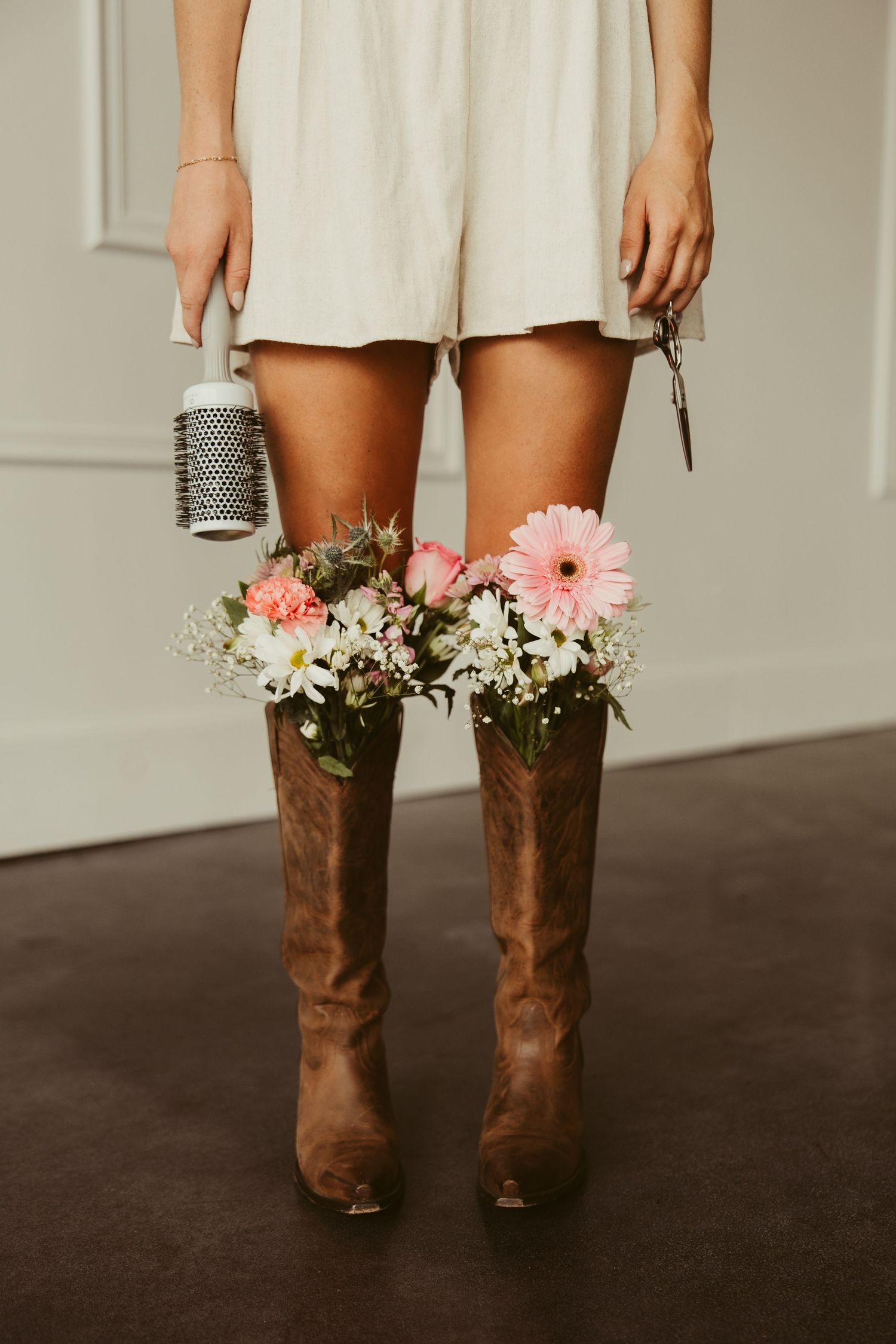 Woman in white dress wearing cowboy boots filled with pink and white spring flowers holding styling tools.
