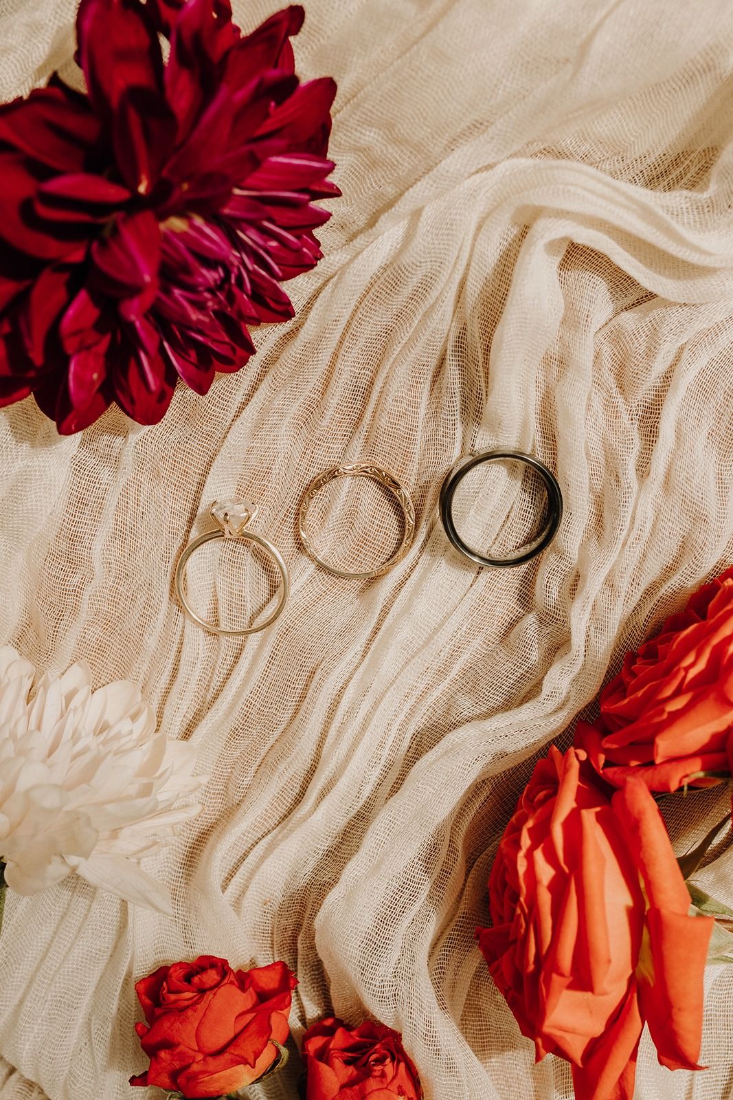 Deep red and coral roses lay artistically arranged on flowing cream-colored fabric with gold wedding rings River Garden Weddings and Events.