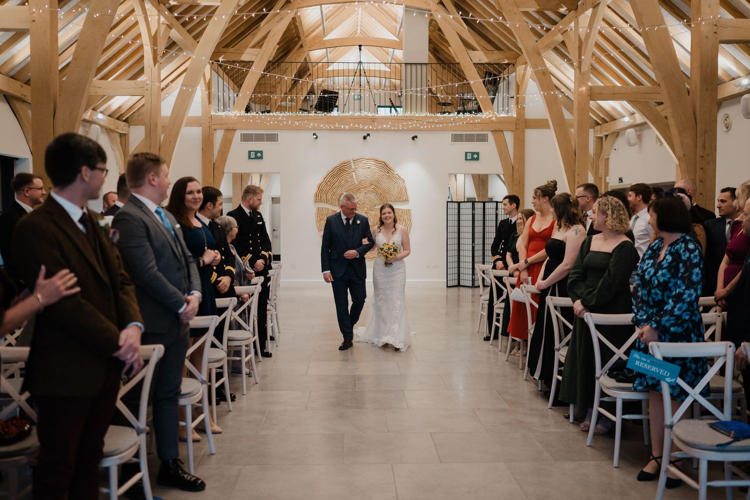 Wedding guests seated in rows of white chairs inside an elegant church venue with wooden beams and white walls.
