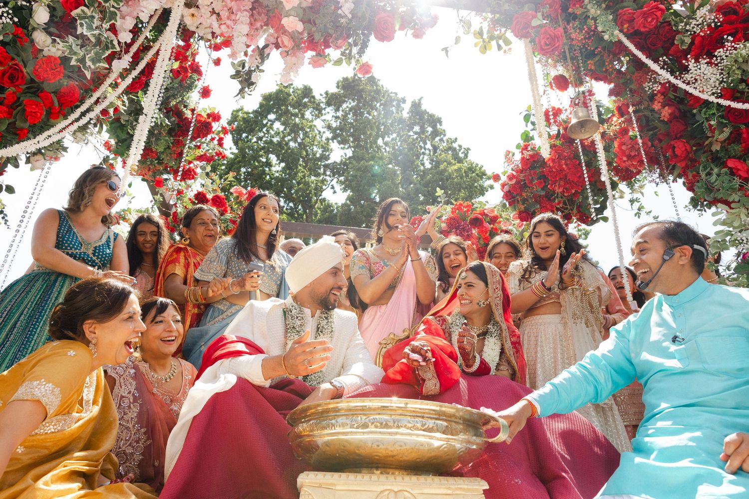 A joyful candid moment of a bride and groom surrounded by laughing family and friends under a lush floral mandap at Ditton Manor, London