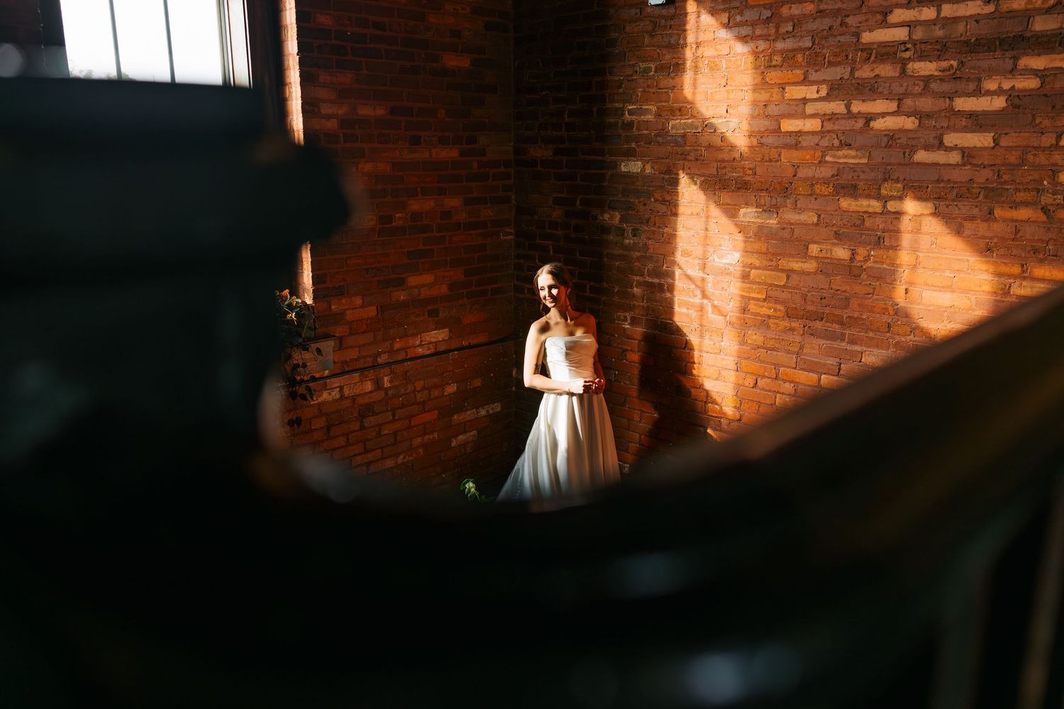 A bride in a white gown stands in dramatic golden light against a rustic brick wall interior.