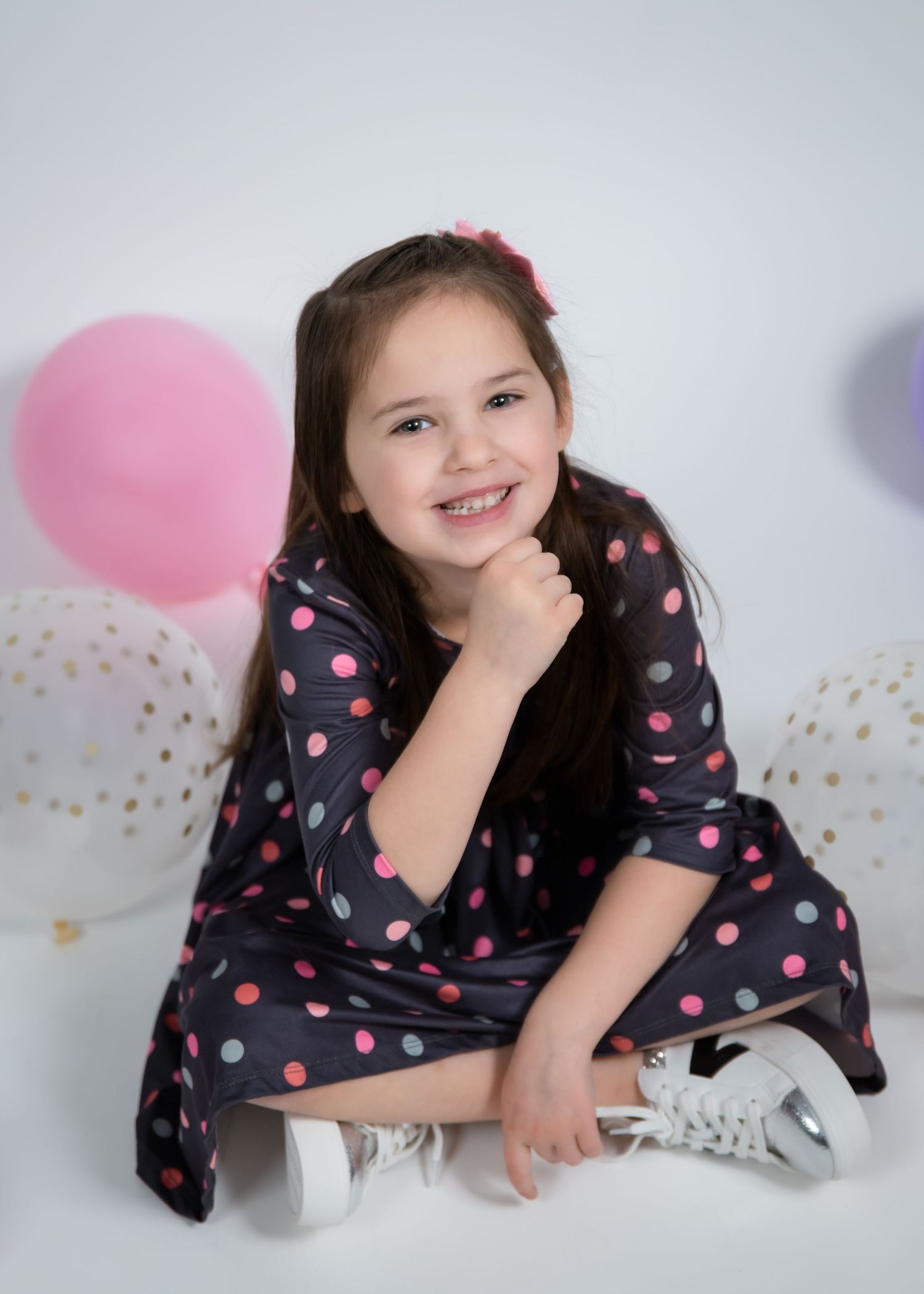 A child in a navy polka dot dress lies playfully on a white floor with pink balloons and gold confetti decorations.