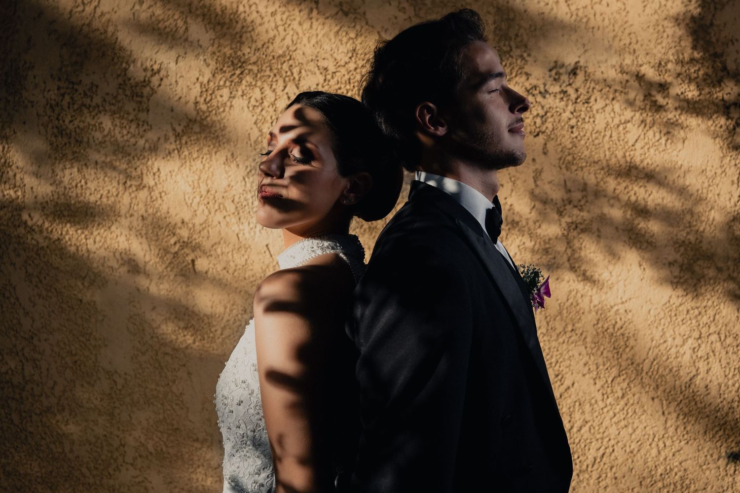 A bride and groom stand back to back in dramatic lighting against a textured wall creating artistic shadows.