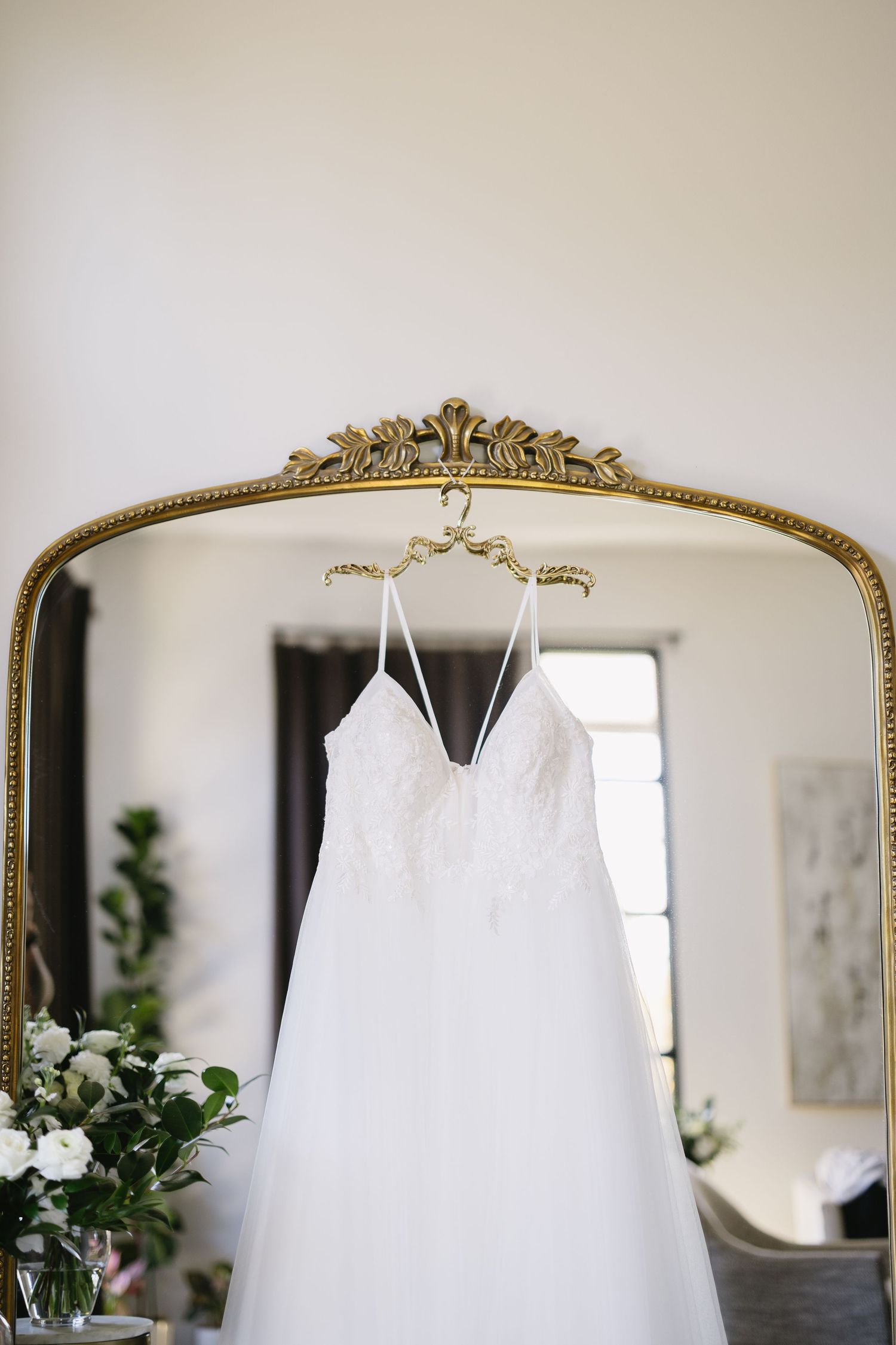 Elegant white wedding dress hanging from an ornate gold mirror frame against a neutral wall.