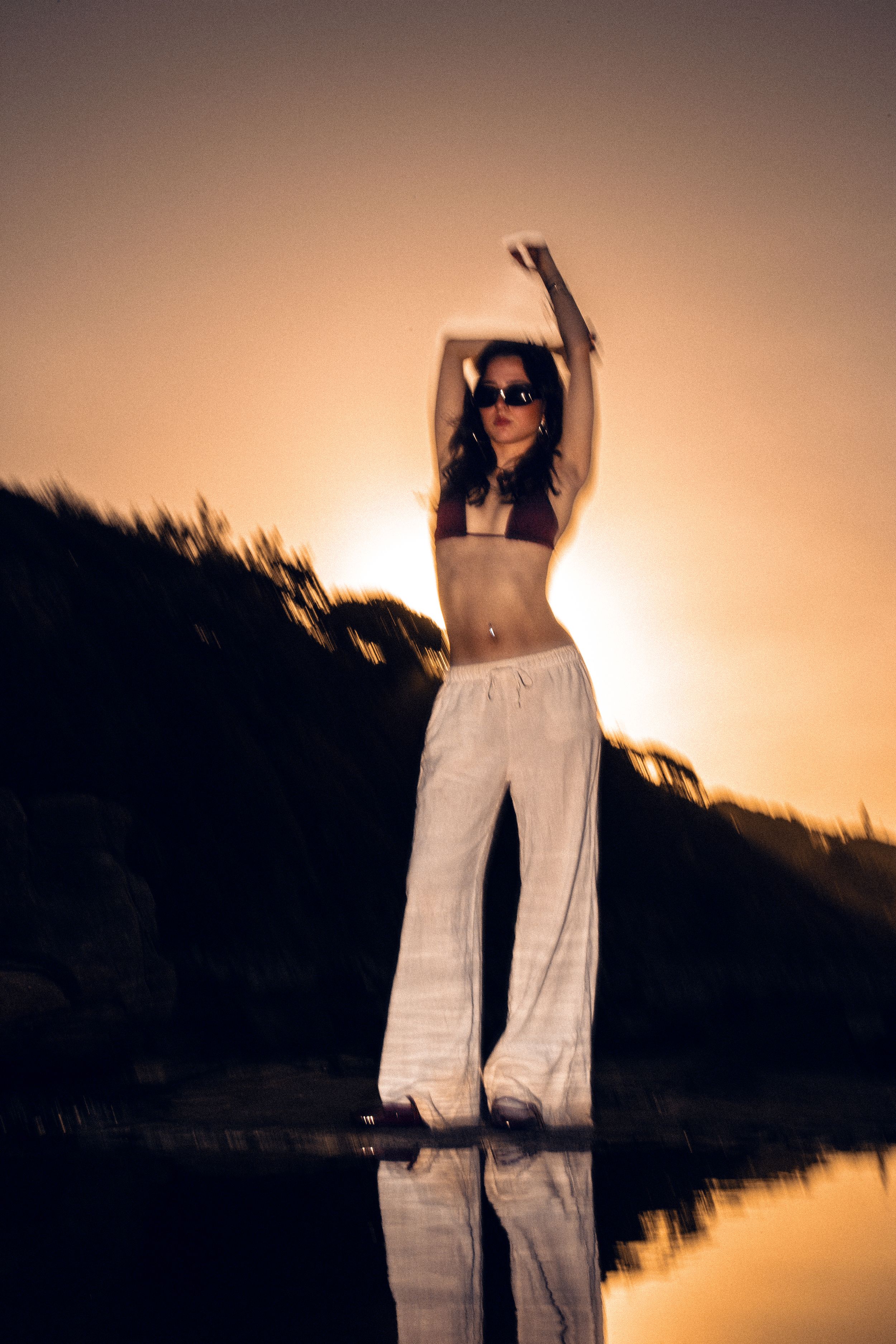 Silhouetted figure in flowing white pants against golden sunset by water.