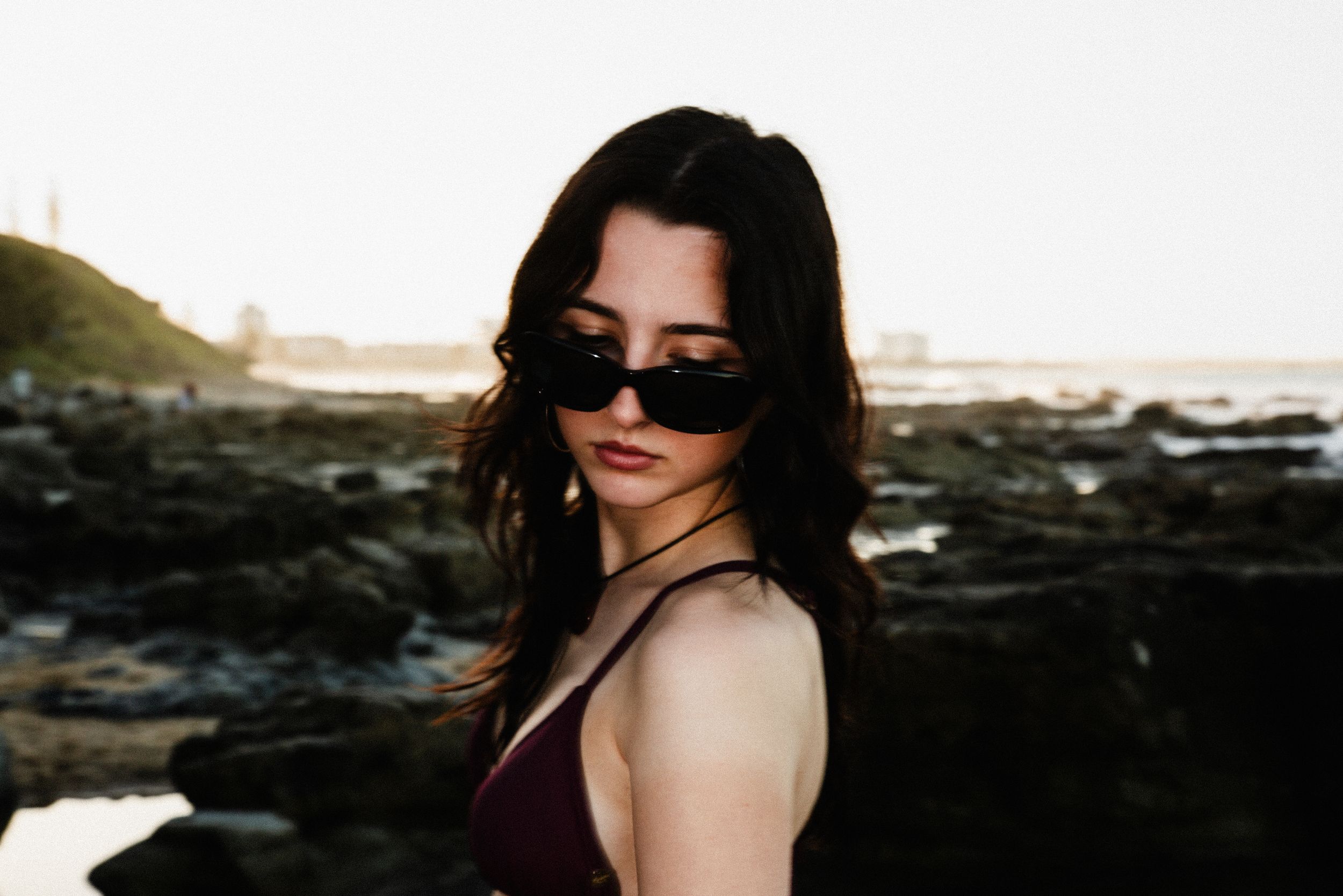 Beachside fashion portraits with dark sunglasses against rocky coastal landscape.