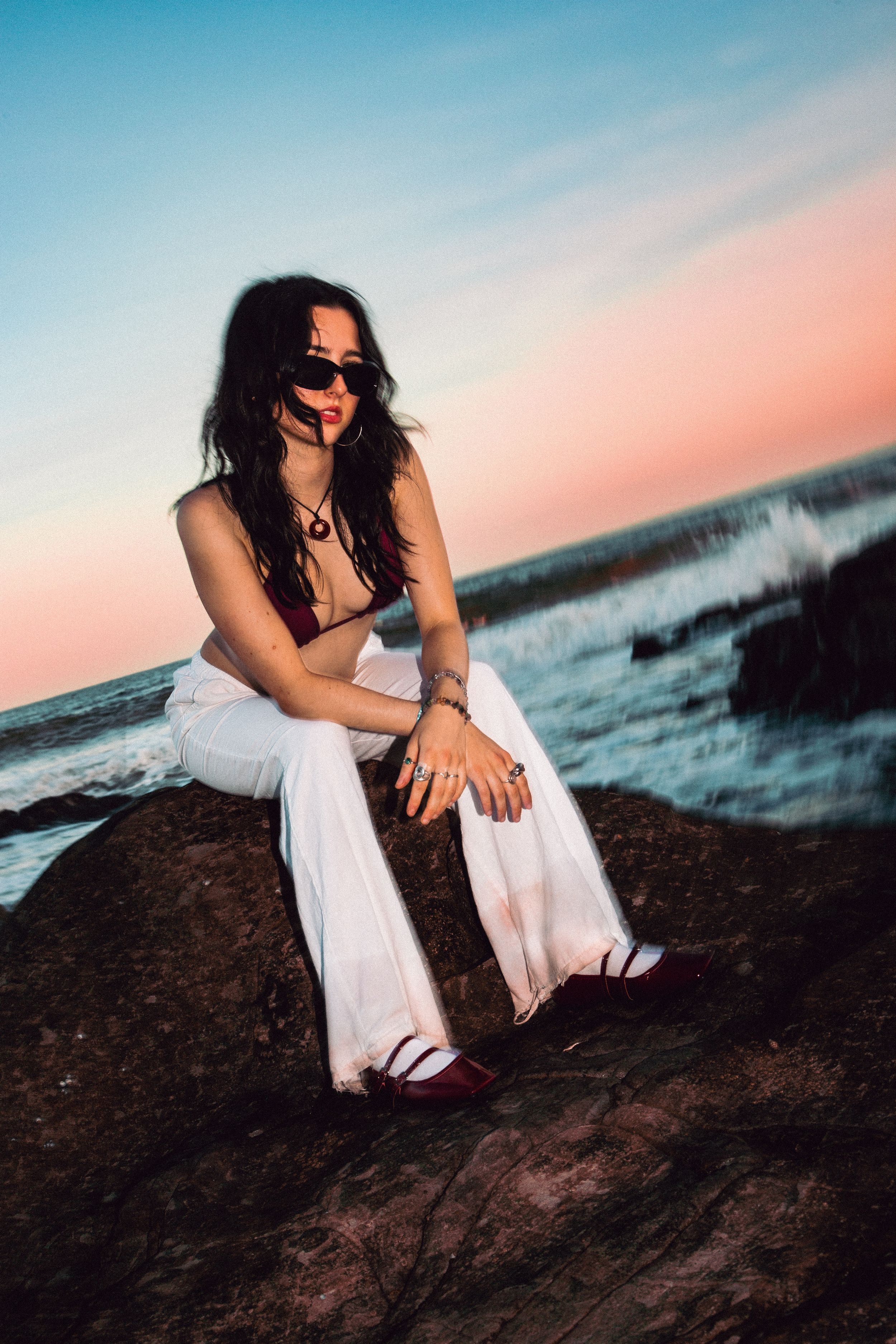 Fashion model in white clothing poses on dark rocks by ocean at twilight.